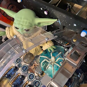May include: A green creature figurine with large ears and a tan robe positioned on a pinball machine playfield. Next to the figurine is a translucent blue circular piece with a white spider-like creature embedded inside, surrounded by metal rails.