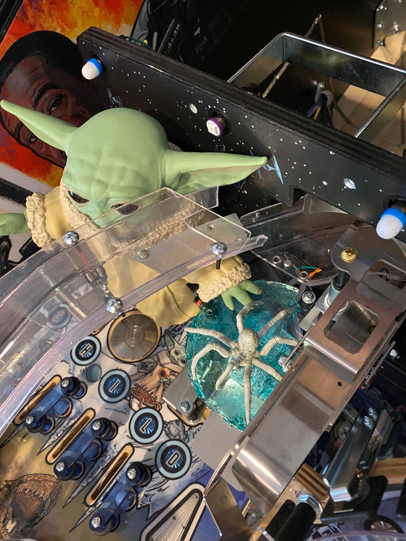 May include: A green creature figurine with large ears and a tan robe positioned on a pinball machine playfield. Next to the figurine is a translucent blue circular piece with a white spider-like creature embedded inside, surrounded by metal rails.