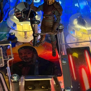 Puede incluir: Primer plano de una m&aacute;quina de pinball con una figura de un personaje blindado sosteniendo un martillo. La m&aacute;quina tiene un dise&ntilde;o colorido y un retrato de un hombre. Luces brillantes iluminan la escena, destacando los intrincados detalles del juego.