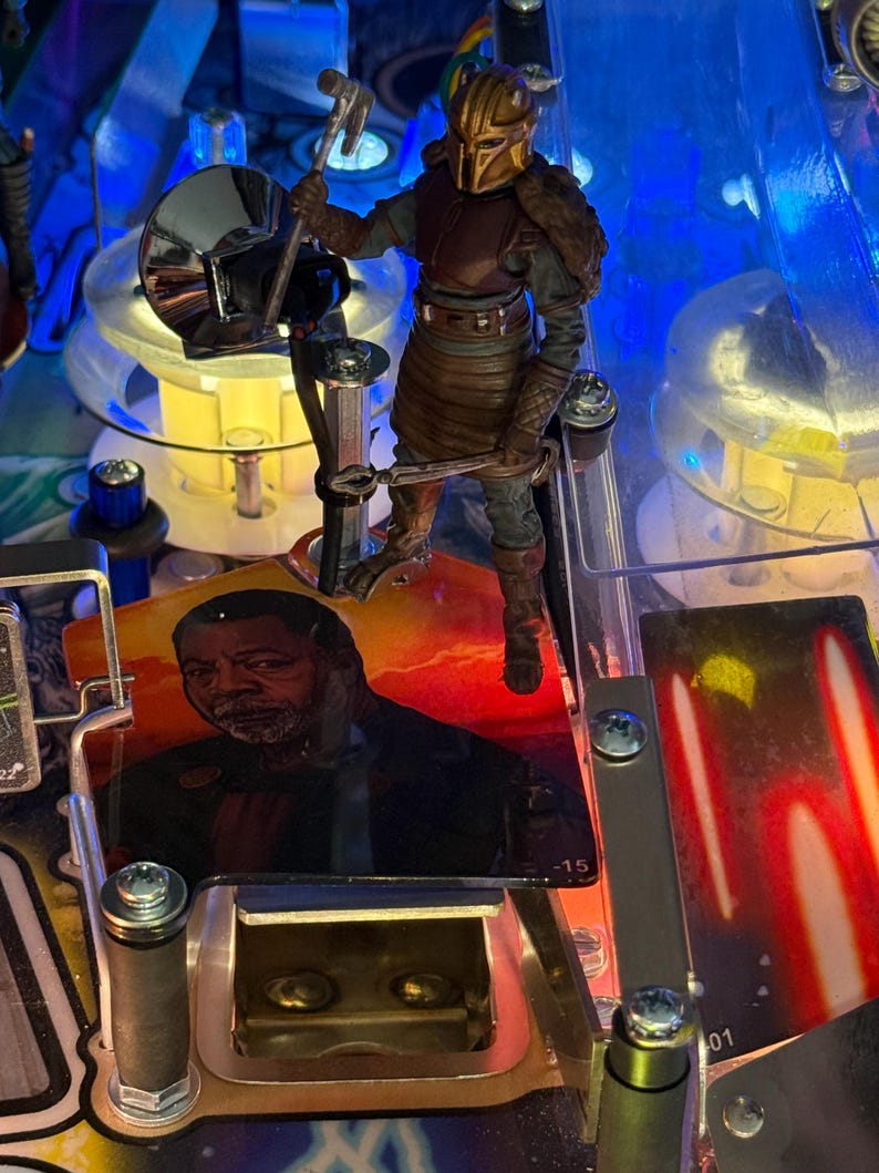 Puede incluir: Primer plano de una m&aacute;quina de pinball con una figura de un personaje blindado sosteniendo un martillo. La m&aacute;quina tiene un dise&ntilde;o colorido y un retrato de un hombre. Luces brillantes iluminan la escena, destacando los intrincados detalles del juego.
