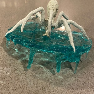 May include: A light gray spider figurine with a textured, pitted body and eight long, segmented legs. The spider is positioned on top of a translucent, bright blue circular base that features dripping, icicle-like resin details along its edges.