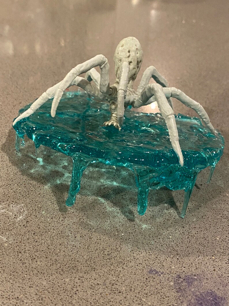 May include: A light gray spider figurine with a textured, pitted body and eight long, segmented legs. The spider is positioned on top of a translucent, bright blue circular base that features dripping, icicle-like resin details along its edges.