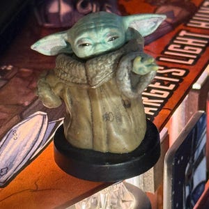 Puede incluir: Una figura de Grogu, tambi&eacute;n conocido como Baby Yoda, de la serie Star Wars. La figura tiene una tez verde claro, orejas grandes y lleva una t&uacute;nica marr&oacute;n. Est&aacute; montada sobre una base negra, con un fondo borroso.