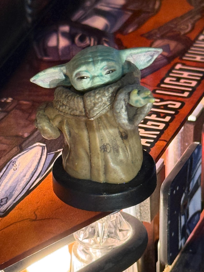 Puede incluir: Una figura de Grogu, tambi&eacute;n conocido como Baby Yoda, de la serie Star Wars. La figura tiene una tez verde claro, orejas grandes y lleva una t&uacute;nica marr&oacute;n. Est&aacute; montada sobre una base negra, con un fondo borroso.
