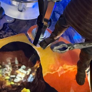 Puede incluir: Primer plano de los componentes internos de una m&aacute;quina de pinball. Se ven varillas met&aacute;licas, una correa negra y una figura con una manga marr&oacute;n. El fondo presenta un dise&ntilde;o abstracto colorido con tonos naranja y amarillo.