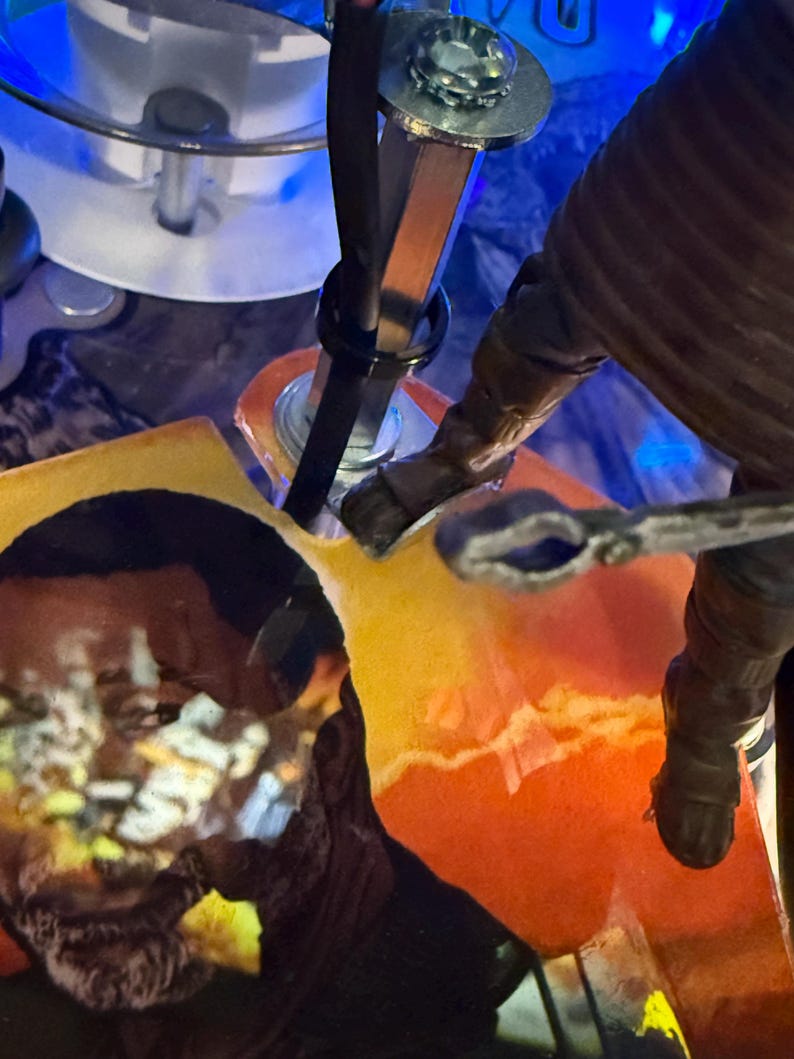 Puede incluir: Primer plano de los componentes internos de una m&aacute;quina de pinball. Se ven varillas met&aacute;licas, una correa negra y una figura con una manga marr&oacute;n. El fondo presenta un dise&ntilde;o abstracto colorido con tonos naranja y amarillo.