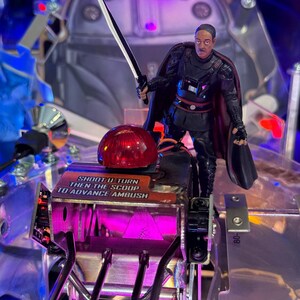 Puede incluir: Una m&aacute;quina de pinball con una figura de un personaje de Star Wars sosteniendo un sable de luz. La m&aacute;quina tiene una c&uacute;pula roja y el texto "SHOOT U-TURN THEN THE SCOOP TO ADVANCE AMBUSH". La m&aacute;quina tiene un esquema de color plateado y p&uacute;rpura.