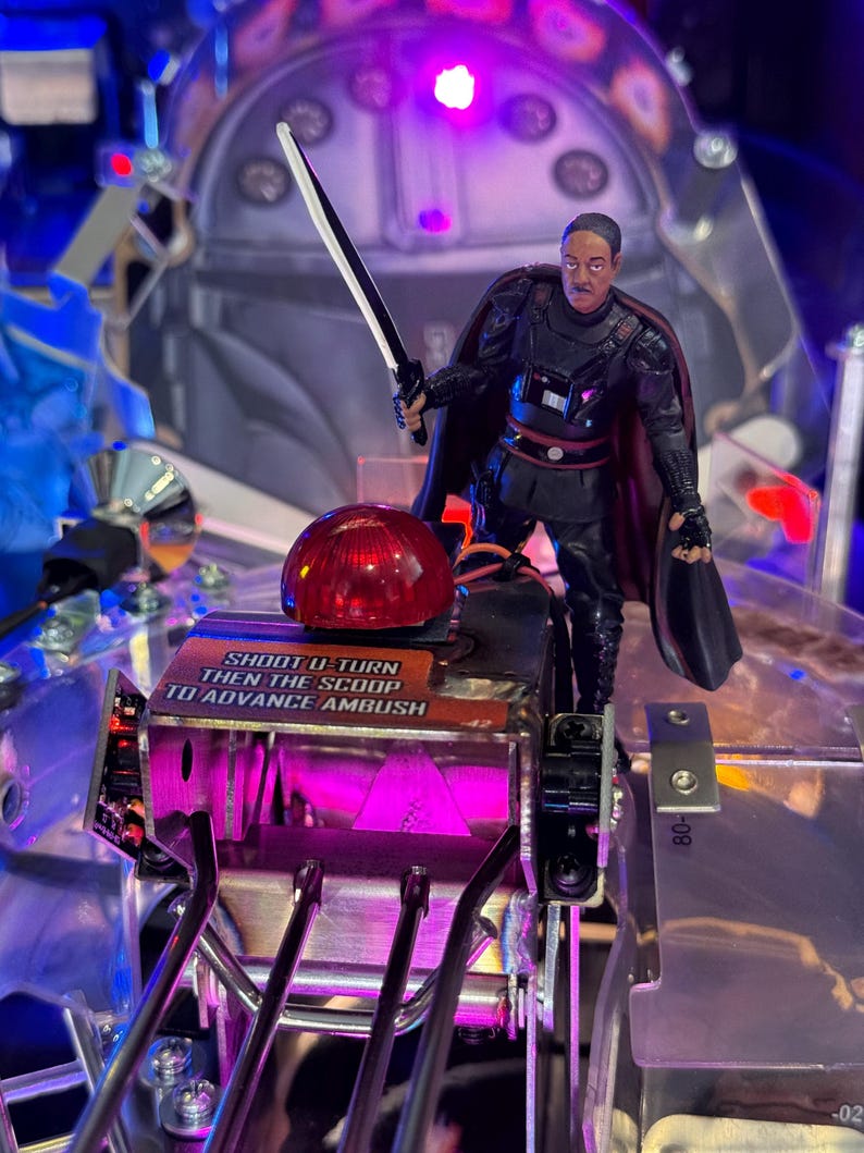 Puede incluir: Una m&aacute;quina de pinball con una figura de un personaje de Star Wars sosteniendo un sable de luz. La m&aacute;quina tiene una c&uacute;pula roja y el texto "SHOOT U-TURN THEN THE SCOOP TO ADVANCE AMBUSH". La m&aacute;quina tiene un esquema de color plateado y p&uacute;rpura.