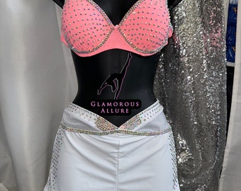 May include: A two-piece dance costume set on a black mannequin. The set includes a pink bedazzled bra and white shorts with rhinestone accents. The bra and shorts are embellished with sparkling rhinestones. The costume is displayed against a silver sequined backdrop.