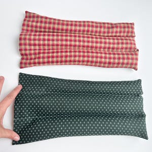 May include: Two rectangular fabric heating pads. The top pad features a red, tan, and dark green plaid pattern. The bottom pad is dark green with a small white geometric diamond print. Both pads are segmented into three horizontal sections.
