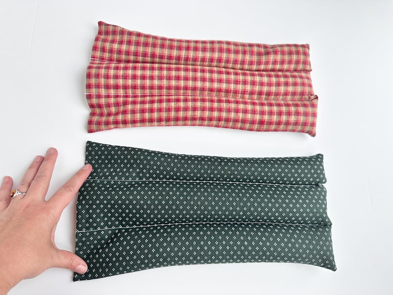 May include: Two rectangular fabric heating pads. The top pad features a red, tan, and dark green plaid pattern. The bottom pad is dark green with a small white geometric diamond print. Both pads are segmented into three horizontal sections.