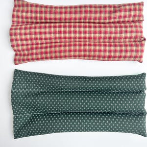 May include: Two rectangular fabric pillows with stitched channels. The top pillow features a red and beige plaid pattern. The bottom pillow is dark green with a repeating pattern of small white four-pointed star shapes across the surface.
