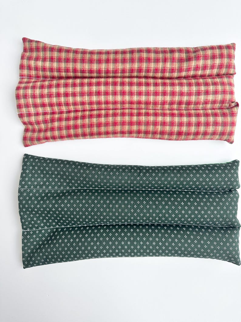 May include: Two rectangular fabric pillows with stitched channels. The top pillow features a red and beige plaid pattern. The bottom pillow is dark green with a repeating pattern of small white four-pointed star shapes across the surface.