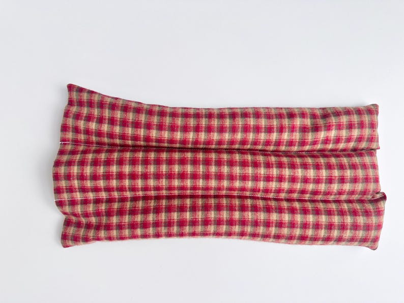 May include: A rectangular fabric heating pad featuring a red and tan plaid pattern. The item is constructed with three horizontal, stitched channels that create a segmented, elongated shape, providing a consistent texture across the surface.