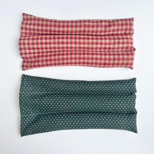 May include: Two rectangular fabric heating pads. The top pad features a red and beige plaid pattern. The bottom pad is dark green with a small white repeating geometric dot pattern. Both pads are segmented into three horizontal sections.