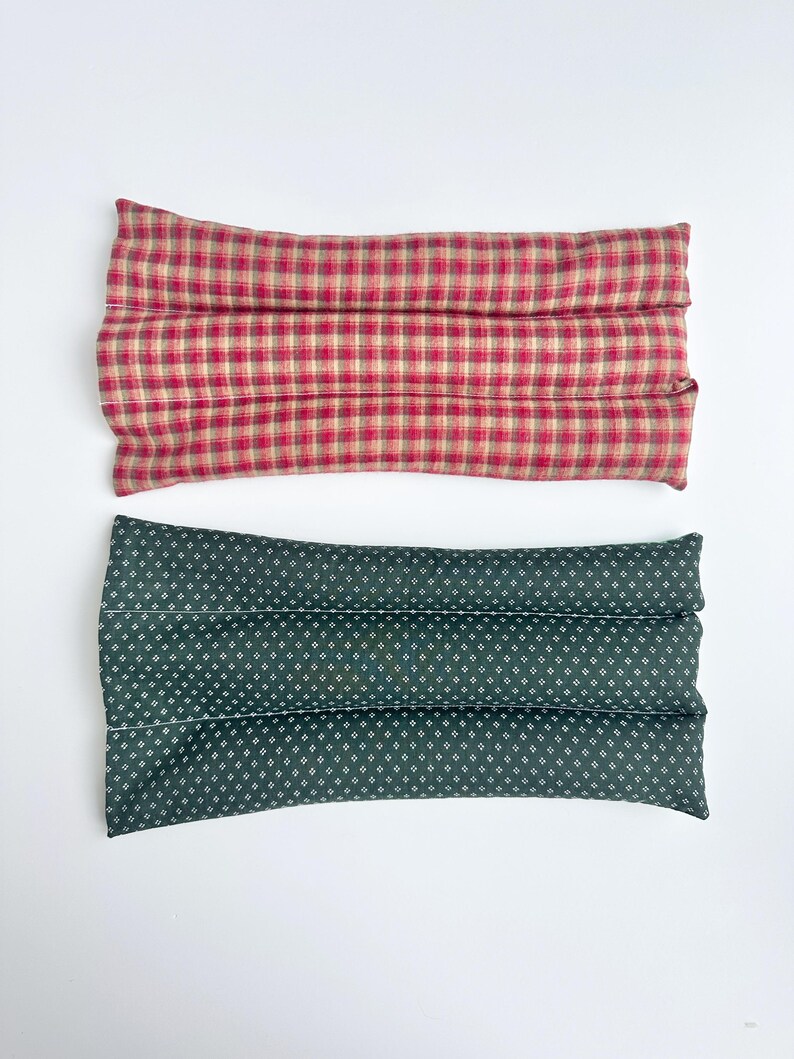 May include: Two rectangular fabric heating pads. The top pad features a red and beige plaid pattern. The bottom pad is dark green with a small white repeating geometric dot pattern. Both pads are segmented into three horizontal sections.