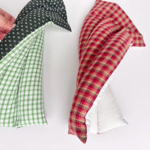 May include: Two rectangular fabric heating pads. One pad features a dark green fabric with a small white geometric pattern on one side and a green and white gingham check on the other. The second pad has a red, brown, and tan plaid pattern.