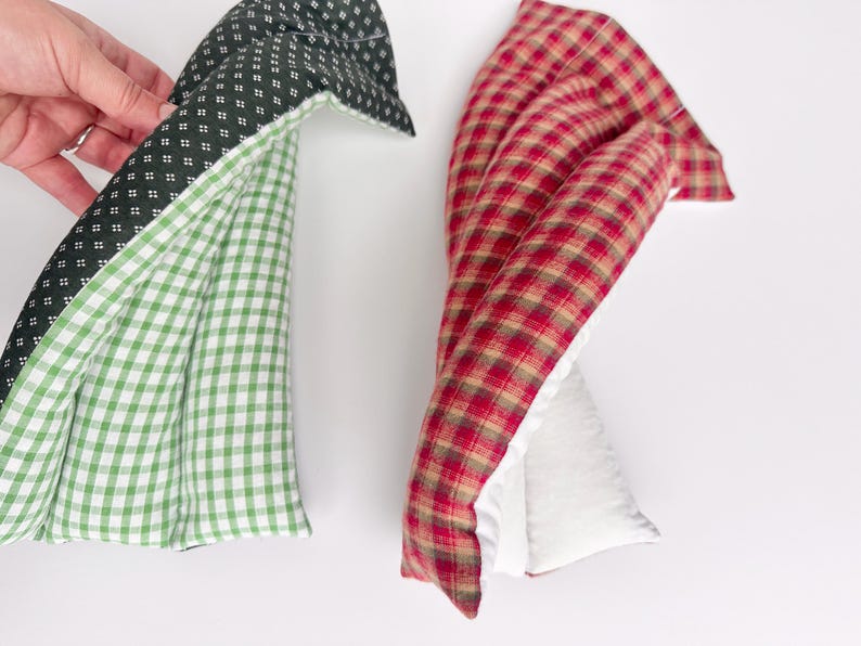 May include: Two rectangular fabric heating pads. One pad features a dark green fabric with a small white geometric pattern on one side and a green and white gingham check on the other. The second pad has a red, brown, and tan plaid pattern.