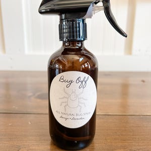 May include: A brown glass bottle with a black spray nozzle and a brown paper label that says "Bug Off" with a cartoon bug and the text "All Natural Bug Spray Lemongrass + Lavender".