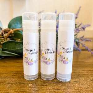 May include: Three lip balm tubes with clear caps and white labels. The labels feature the text "Jemyn & Lavender" in a script font, surrounded by a floral wreath with purple and yellow flowers. The tubes are on a wooden surface.