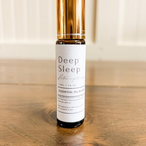 May include: A small essential oil blend bottle with a gold cap and a white label. The label reads "Deep Sleep" and "Essential Oil Blend". The bottle is 10ml / .34 oz. The bottle is on a wooden surface.
