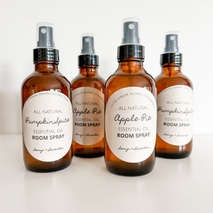 May include: Four amber glass bottles of essential oil room spray. Each bottle has a black spray nozzle and a white label with the scent name: Pumpkin Spice and Apple Pie. The labels also include the words "All Natural" and "Essential Oil Room Spray."