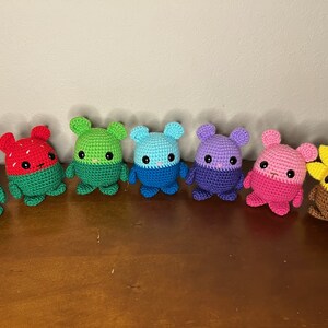 May include: A row of seven crocheted stuffed toys in various colors, including pink, red, green, blue, purple, and yellow. The toys are all in the shape of a small animal with a round body and a small head. The toys are all smiling and have big eyes.