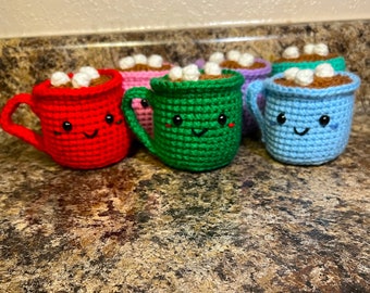 May include: Five crocheted mugs with smiling faces, each filled with white marshmallows. The mugs are red, green, pink, blue, and light blue.