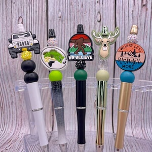 May include: Five different pens with colorful, themed toppers. The toppers include a jeep, a fish, a Bigfoot, a deer, and a mountain scene with the text "Worry Less Wander More".