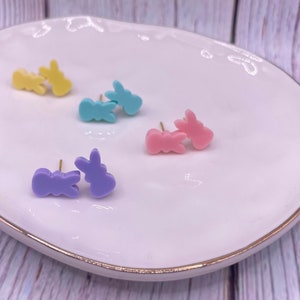 May include: Four pairs of bunny-shaped stud earrings in pastel colors: yellow, blue, purple, and pink. The earrings are displayed on a white plate with a gold rim.
