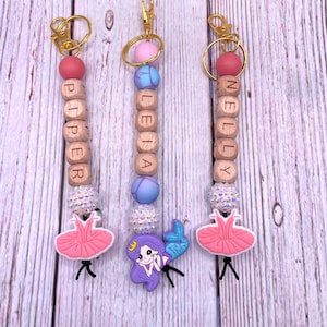 May include: Three personalized wooden bead keychains with a pink, blue, and pink silicone charm. Each keychain features a different name: Piper, Leia, and Nelly. The charms are a ballerina, a mermaid, and a ballerina.