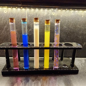 May include: Six glass test tubes filled with different colored liquids are arranged in a black test tube rack. The liquids are purple, blue, white, yellow, and two shades of purple.