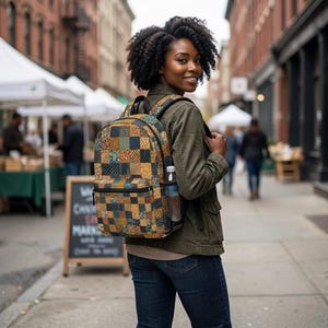 May include: A backpack featuring a multi-colored patchwork pattern of squares in shades of mustard yellow, dark blue, and brown. The bag includes a front zippered pocket, a top handle, adjustable shoulder straps, and a side mesh water bottle pocket.