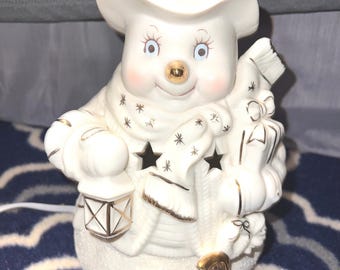 May include: A ceramic snowman figurine with a top hat, scarf, and holding a lantern and gifts. The snowman is white with gold accents, rosy cheeks, and blue eyes. The scarf has star cutouts.
