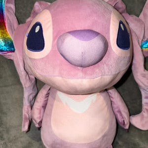 Puede incluir: Un mu&ntilde;eco de peluche rosa de Angel con ojos grandes de color azul oscuro y una nariz morada. El mu&ntilde;eco tiene orejas grandes y un dise&ntilde;o blanco en el pecho. Las puntas de las orejas tienen un dise&ntilde;o de arco&iacute;ris.