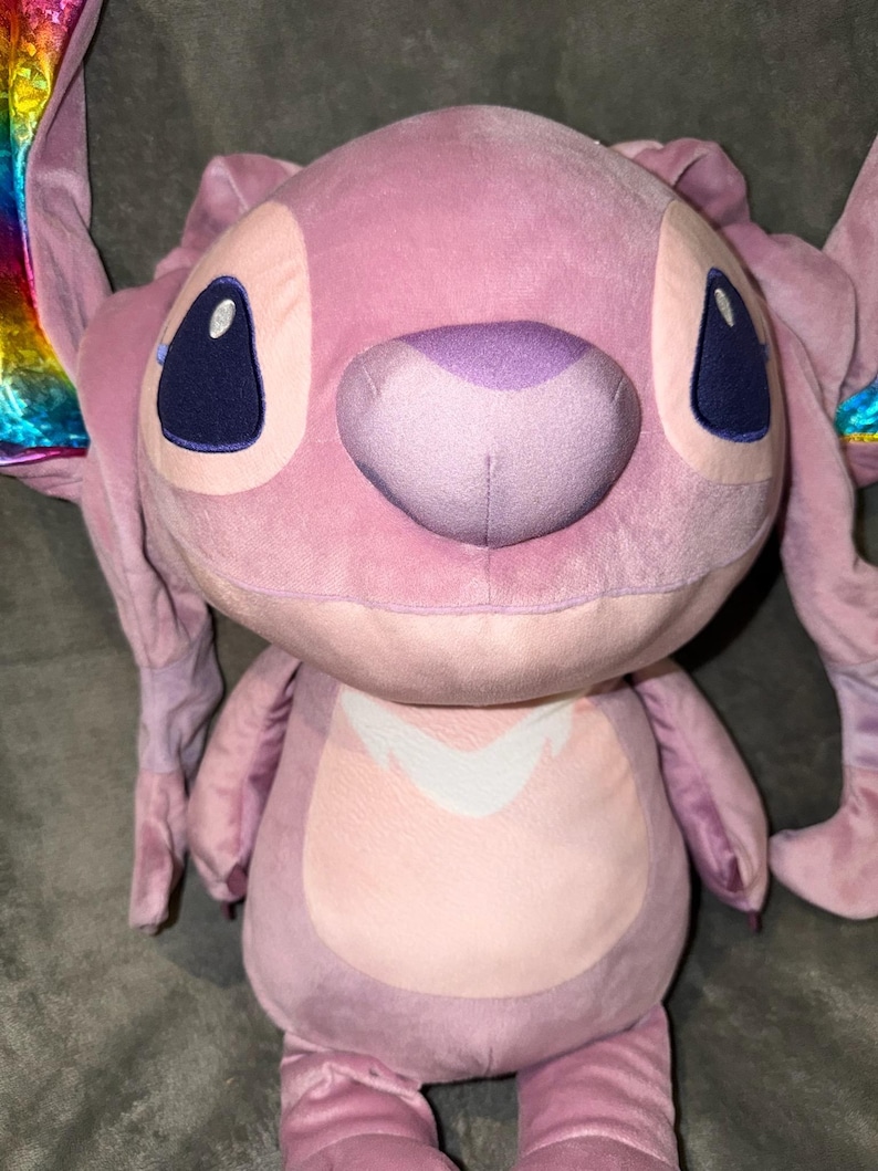 Puede incluir: Un mu&ntilde;eco de peluche rosa de Angel con ojos grandes de color azul oscuro y una nariz morada. El mu&ntilde;eco tiene orejas grandes y un dise&ntilde;o blanco en el pecho. Las puntas de las orejas tienen un dise&ntilde;o de arco&iacute;ris.