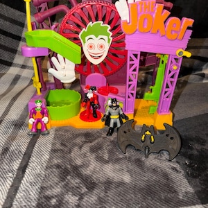 May include: A colorful Joker-themed amusement park playset featuring a large red Ferris wheel, a green slide, and a purple sign that reads THE JOKER. Included are three small action figures of the Joker, Harley Quinn, and Batman, plus a black bat-shaped accessory.