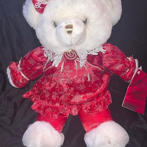 May include: White plush teddy bear wearing a red velvet dress with gold trim and a white lace collar. The bear is wearing a white fur hat with a gold bow and a gold swirl design. The bear is sitting on a black background. The tag reads "Keepsake Holiday Bear".