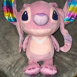 Puede incluir: Un peluche rosa de un personaje alien&iacute;gena con grandes orejas con un brillo met&aacute;lico arco&iacute;ris. El juguete tiene ojos grandes de color azul oscuro, una nariz p&uacute;rpura y una barriga rosa claro. El peluche est&aacute; sobre una superficie gris.