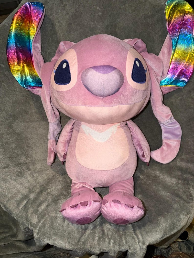 Puede incluir: Un peluche rosa de un personaje alien&iacute;gena con grandes orejas con un brillo met&aacute;lico arco&iacute;ris. El juguete tiene ojos grandes de color azul oscuro, una nariz p&uacute;rpura y una barriga rosa claro. El peluche est&aacute; sobre una superficie gris.