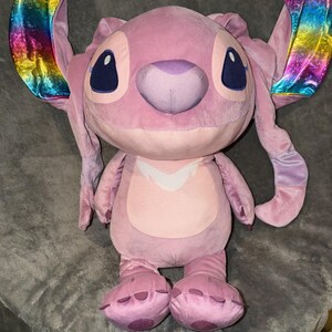 Puede incluir: Un personaje de peluche rosa con grandes orejas de colores del arco&iacute;ris. El peluche tiene ojos grandes de color azul oscuro, una nariz p&uacute;rpura y una barriga rosa claro. El juguete est&aacute; sobre una superficie gris.