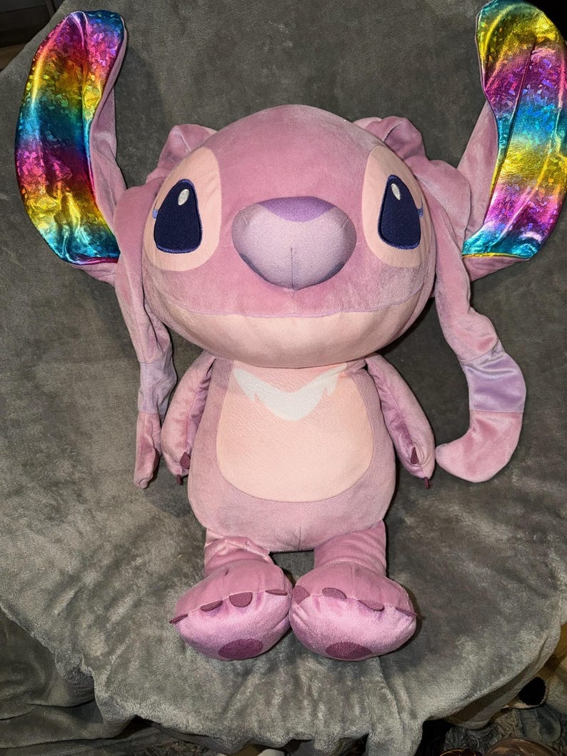 Puede incluir: Un personaje de peluche rosa con grandes orejas de colores del arco&iacute;ris. El peluche tiene ojos grandes de color azul oscuro, una nariz p&uacute;rpura y una barriga rosa claro. El juguete est&aacute; sobre una superficie gris.