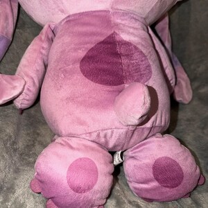 Puede incluir: Un peluche rosa y morado, con un parche en forma de coraz&oacute;n en la espalda y grandes pies redondeados con c&iacute;rculos rosas m&aacute;s oscuros. El juguete tiene una textura suave y difusa y se asemeja a un personaje de dibujos animados.