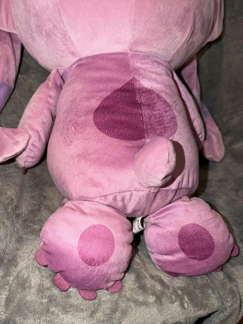 Puede incluir: Un peluche rosa y morado, con un parche en forma de coraz&oacute;n en la espalda y grandes pies redondeados con c&iacute;rculos rosas m&aacute;s oscuros. El juguete tiene una textura suave y difusa y se asemeja a un personaje de dibujos animados.