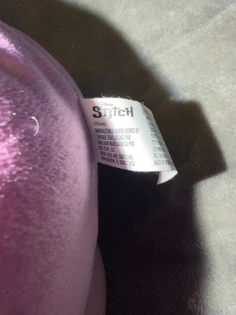 Puede incluir: Primer plano de un peluche con una etiqueta. La etiqueta dice "Stitch" y "&copy;Disney". La etiqueta tambi&eacute;n incluye informaci&oacute;n de fabricaci&oacute;n y la direcci&oacute;n de Just Play, LLC en Boca Raton, Florida.
