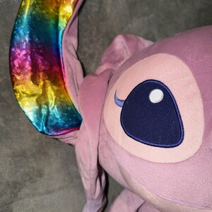 Puede incluir: Un peluche rosa con la cara de un personaje y orejas grandes. Una oreja es un vibrante arco&iacute;ris de colores, la otra es rosa. El personaje tiene un ojo grande, azul oscuro con un reflejo blanco y una ceja peque&ntilde;a y curvada.