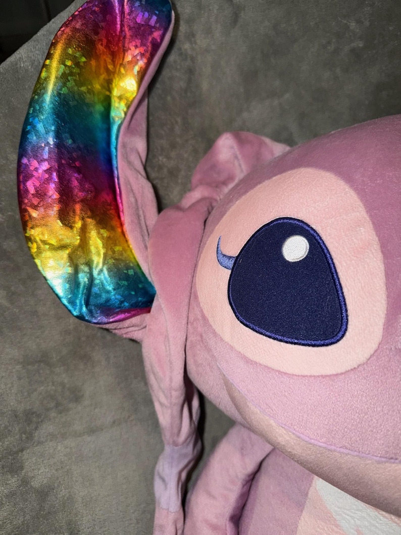 Puede incluir: Un peluche rosa con la cara de un personaje y orejas grandes. Una oreja es un vibrante arco&iacute;ris de colores, la otra es rosa. El personaje tiene un ojo grande, azul oscuro con un reflejo blanco y una ceja peque&ntilde;a y curvada.