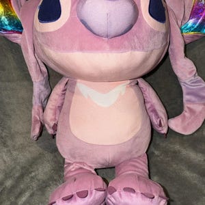 Puede incluir: Un peluche rosa del personaje Angel de Lilo & Stitch. El juguete tiene detalles en rosa oscuro en los pies y las orejas, ojos azules grandes y un vientre rosa claro. Las alas tienen un brillo iridiscente arco&iacute;ris.