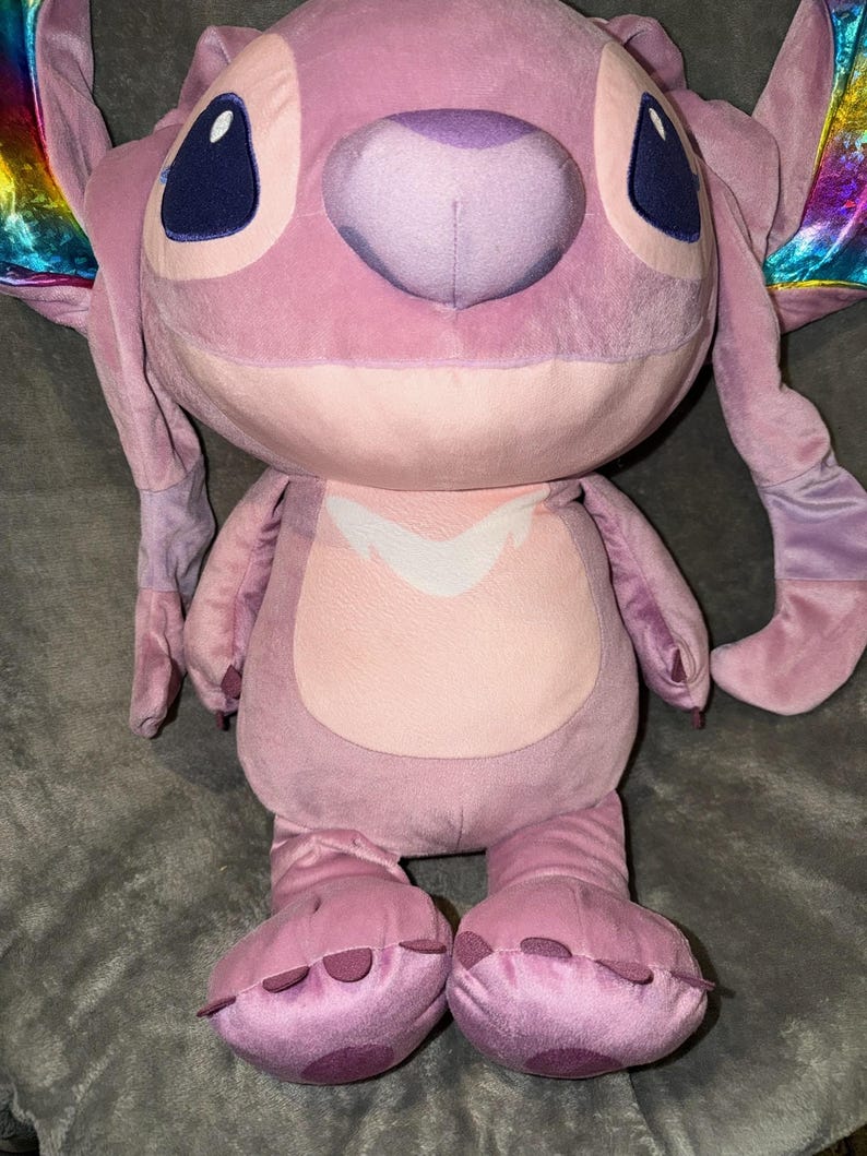 Puede incluir: Un peluche rosa del personaje Angel de Lilo & Stitch. El juguete tiene detalles en rosa oscuro en los pies y las orejas, ojos azules grandes y un vientre rosa claro. Las alas tienen un brillo iridiscente arco&iacute;ris.