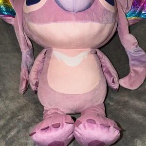 Puede incluir: Una gran mu&ntilde;eca de peluche Angel en tonos rosas y morados. La mu&ntilde;eca tiene ojos azules grandes, orejas largas y un hocico redondeado. Los pies de la mu&ntilde;eca tienen manchas moradas oscuras. La mu&ntilde;eca tiene alas de colores arco&iacute;ris.